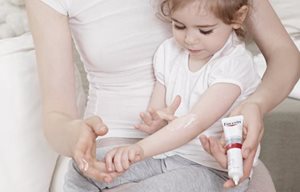 Liten flicka som sitter i sin mors knä och applicerar Eucerin AtoControl Acute Care Cream i armvecket.